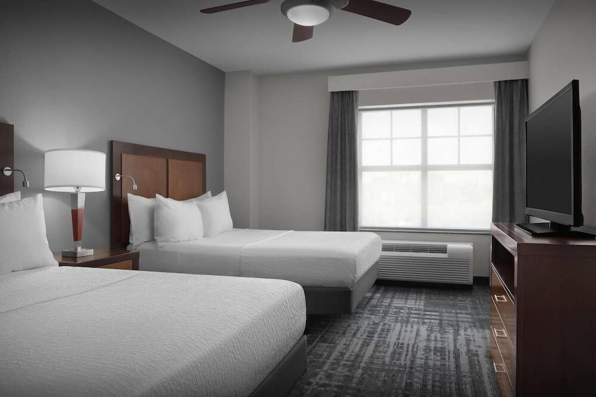 Ein Hotelzimmer mit zwei Betten, einem Deckenventilator, einem Fernseher und einem großen Fenster mit Vorhängen.