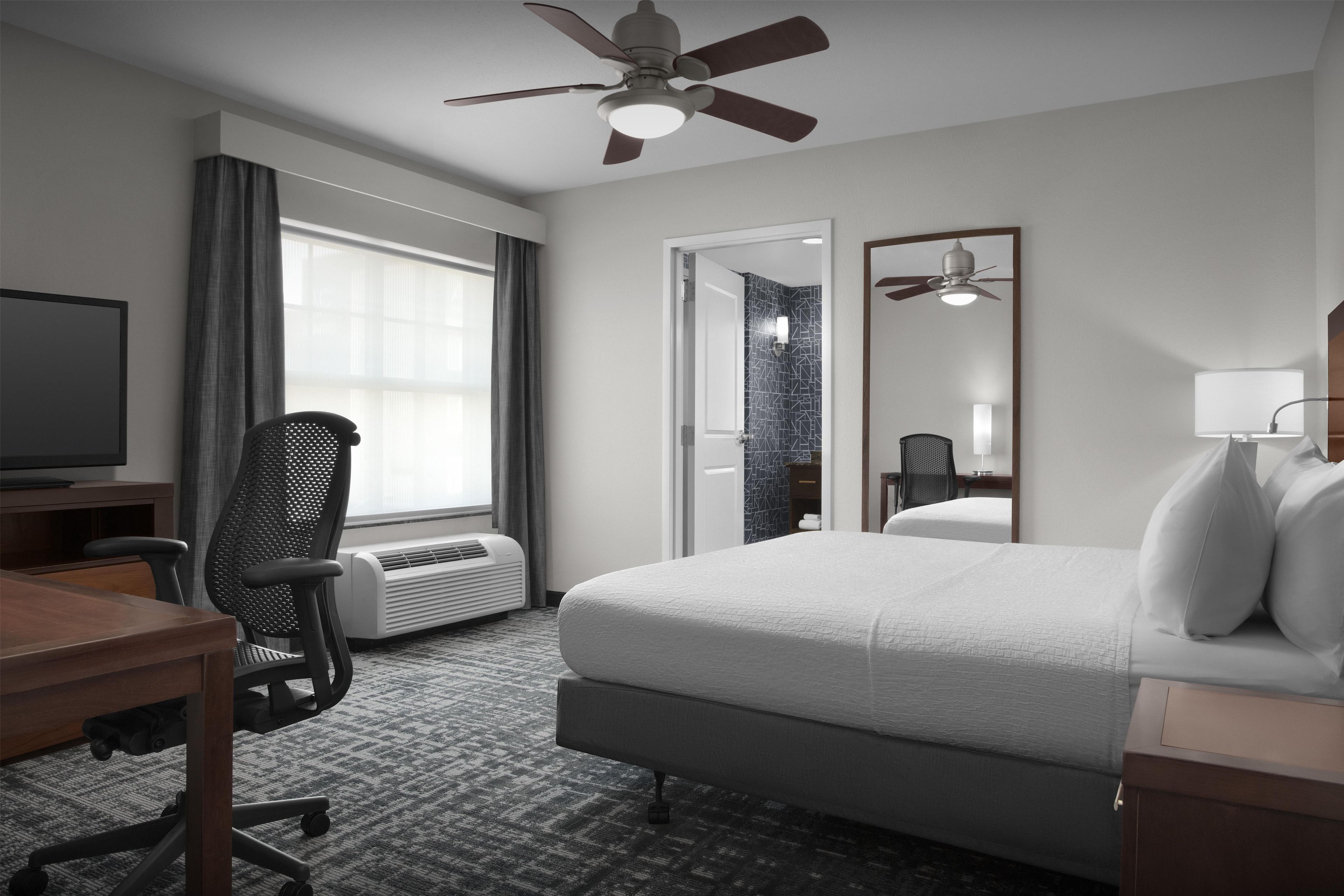 A hotel room with a bed, desk, chair, television, ceiling fan, and a large window with curtains.