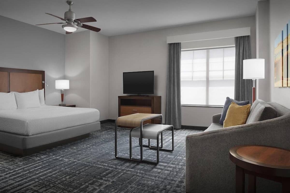 Ein Hotelzimmer mit Bett, Fernseher, Sofa, kleinem Tisch, Stuhl, Deckenventilator und einem Fenster mit Vorhängen.