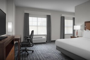 Habitación de hotel con cama, escritorio con silla, televisor, lámpara y ventana con cortinas.