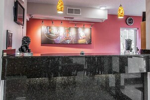 Una zona de recepción con mostrador de mármol negro, un televisor de pared grande que muestra un evento musical, y un reloj encima.