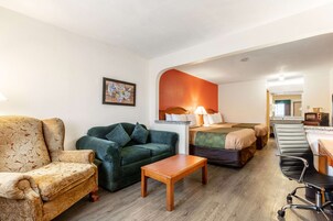 Habitación de hotel con sofá, un sillón verde, una mesa de madera, cama con cubrecama verde, una silla de oficina negra y una pequeña kitchenette.