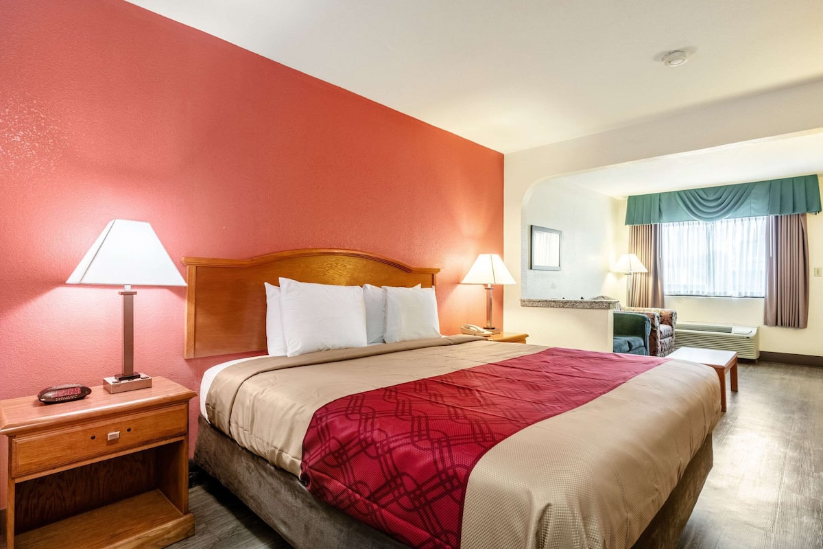 A hotel room with a bed, two bedside tables with lamps, a nightstand, a window with curtains, and a wall-mounted air conditioning unit.