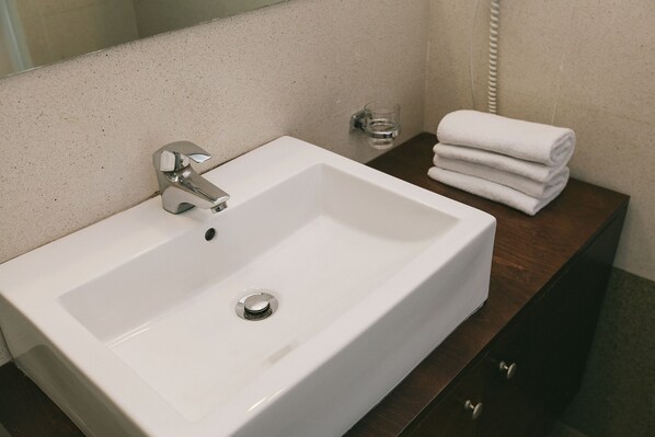 A white bathroom sink with a chrome faucet, a glass of water, and a stack of white towels on a wooden counter. - Du Lac Hotel (Agios Nikolaos)