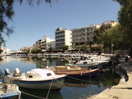 Una marina con barcos amarrados, un hotel llamado "Hotel Du Lac" al fondo y un paseo marítimo con árboles y edificios.