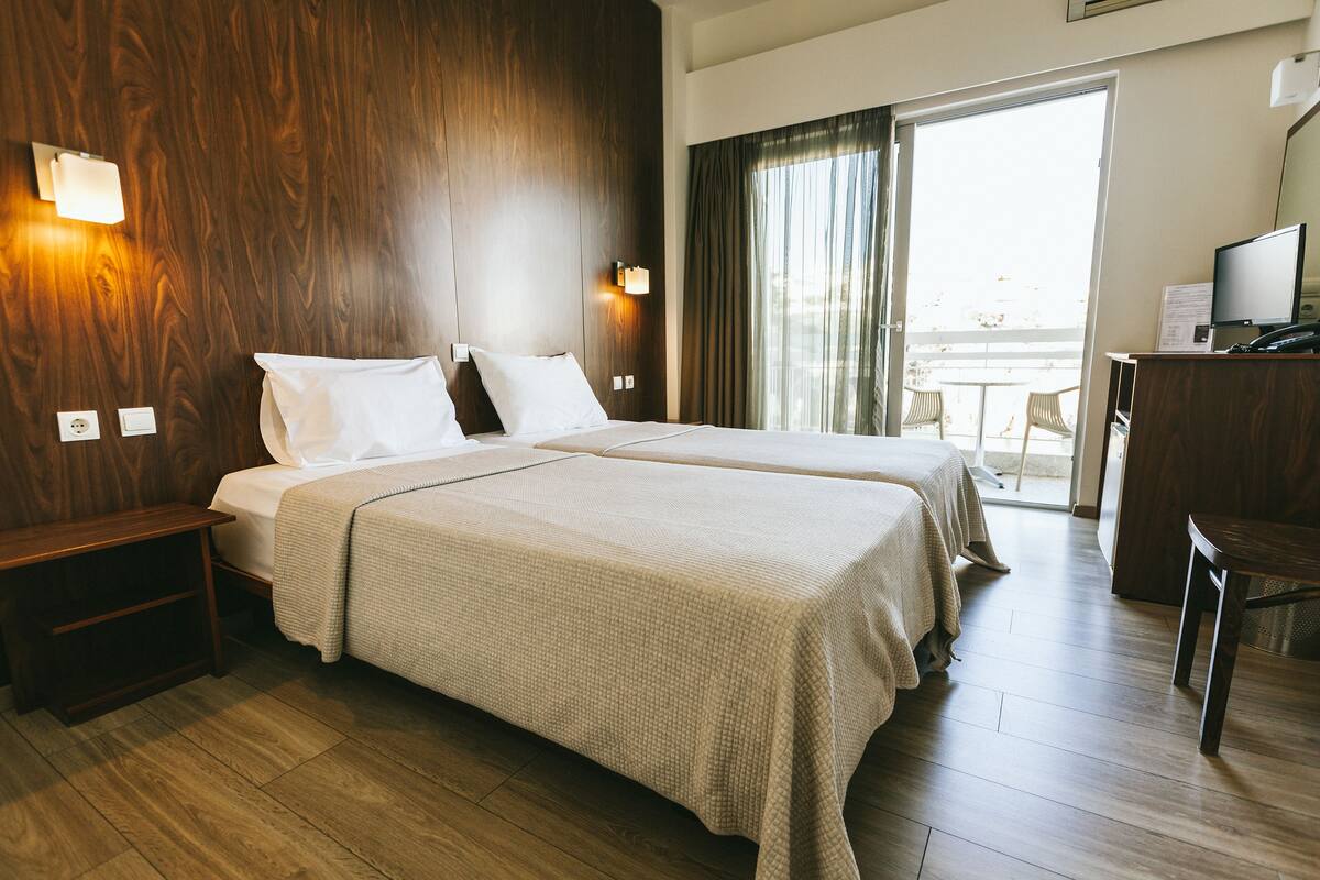 A hotel room with a wooden floor, a bed with white linens, a desk with a television, and a view of the outdoors through a sliding door.