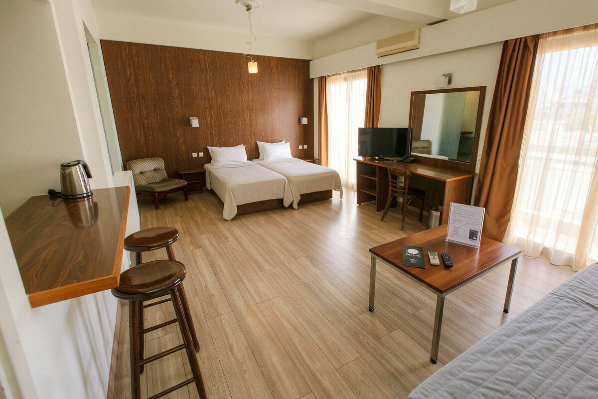 A hotel room with a wooden floor, a bed with two pillows, a desk with a computer and a TV, a coffee maker, and a mirror on the wall.