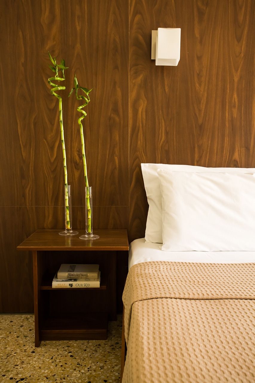 Habitación de hotel con pared de madera, una mesita de noche con un libro, una cama con almohada blanca y dos plantas de bambú en jarrones de vidrio.