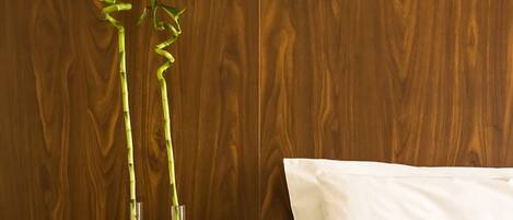 Habitación de hotel con pared de madera, una mesita de noche con un libro, una cama con almohada blanca y dos plantas de bambú en jarrones de vidrio.