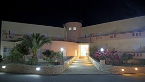 A hotel entrance with a curved pathway, stone planters with plants, and a central illuminated doorway. - Hotel Sea Breeze (Sitia)