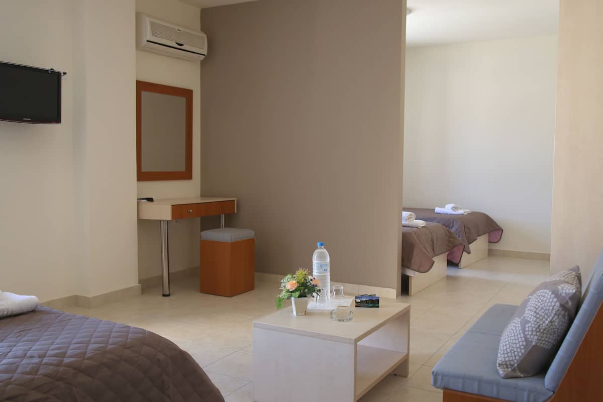 Une chambre d’hôtel avec un lit, un canapé, un bureau avec un miroir, une petite table avec une bouteille d’eau et un vase de fleurs, et un deuxième lit à l’arrière-plan.