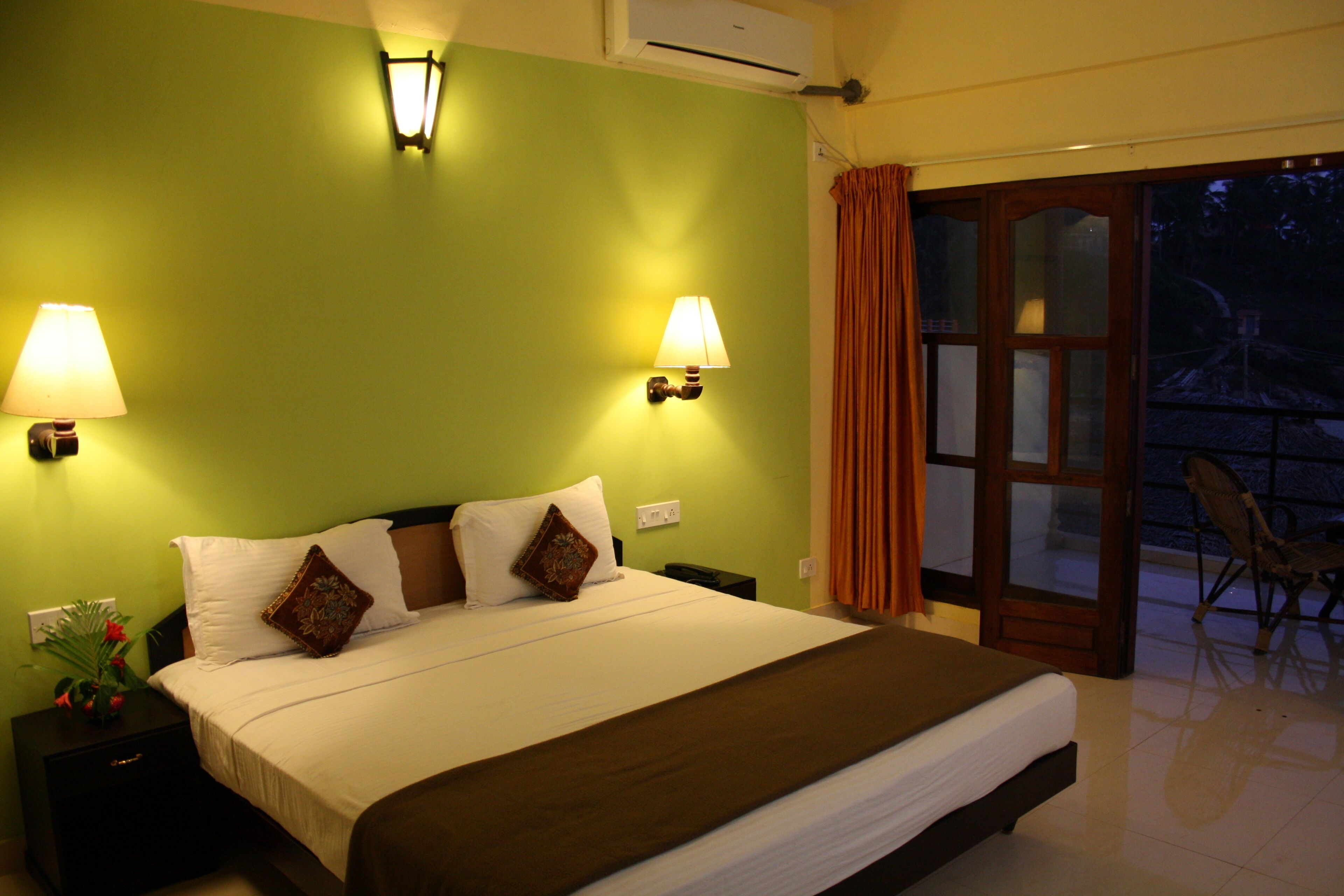 Habitación de hotel con una cama grande, dos lámparas de pared, una mesita de noche con una planta y vistas al exterior a través de una puerta de cristal.