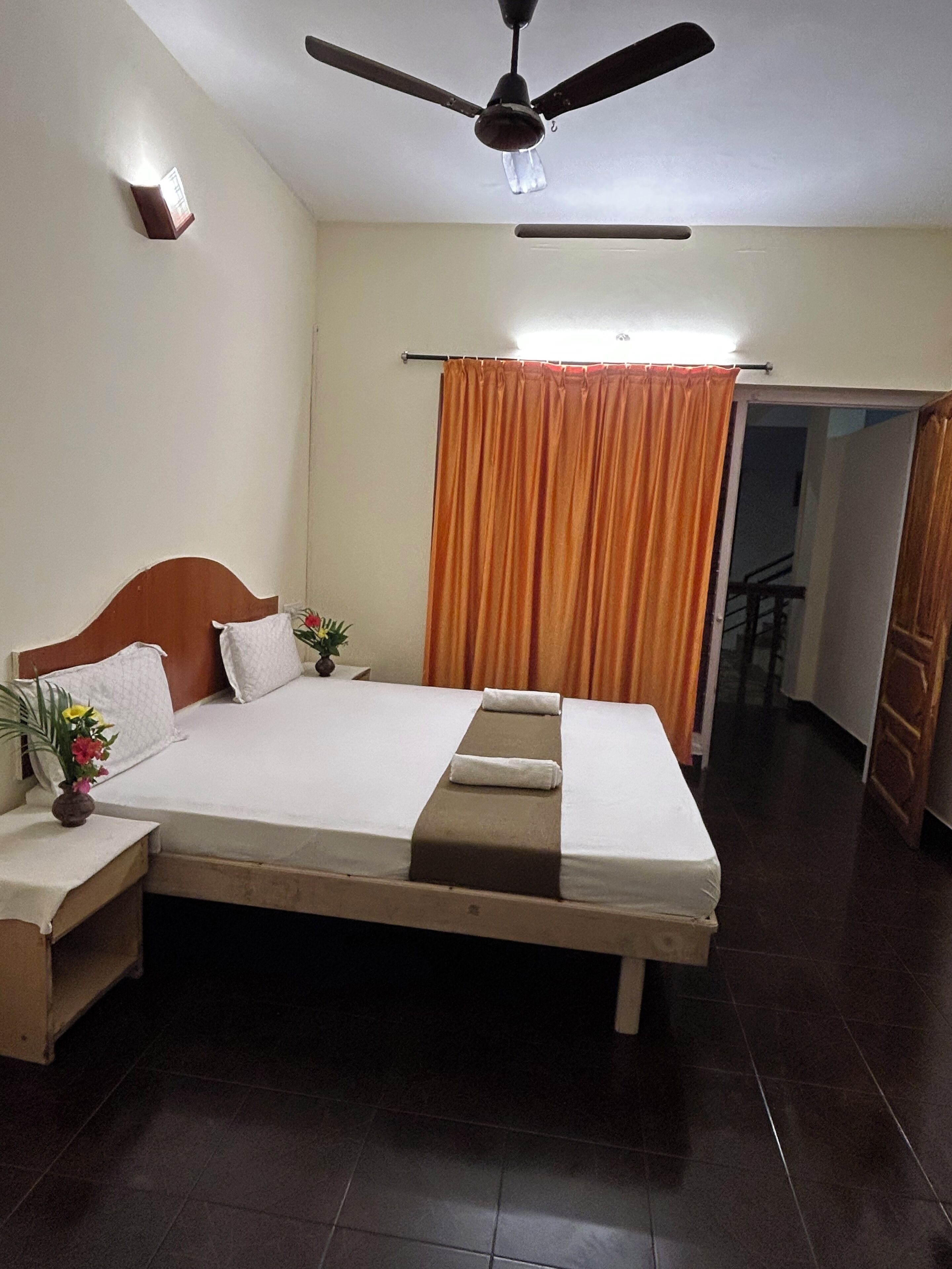 Habitación de hotel con cama, mesita de noche, ventilador de techo, ventana con cortinas y puerta que da a otra habitación.