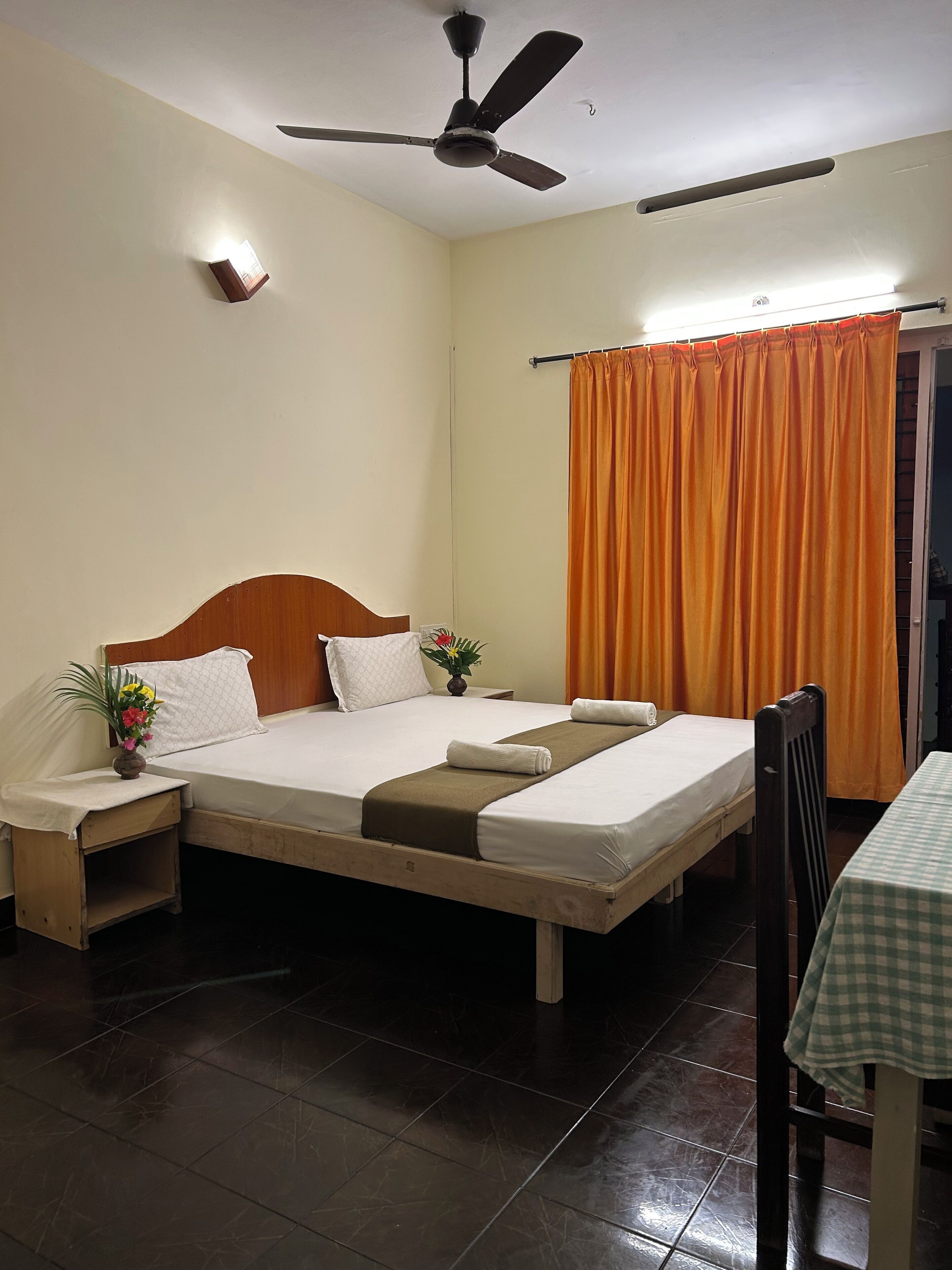 A neatly arranged hotel room with a bed, bedside tables, a chair, a table, a ceiling fan, and a window with curtains.