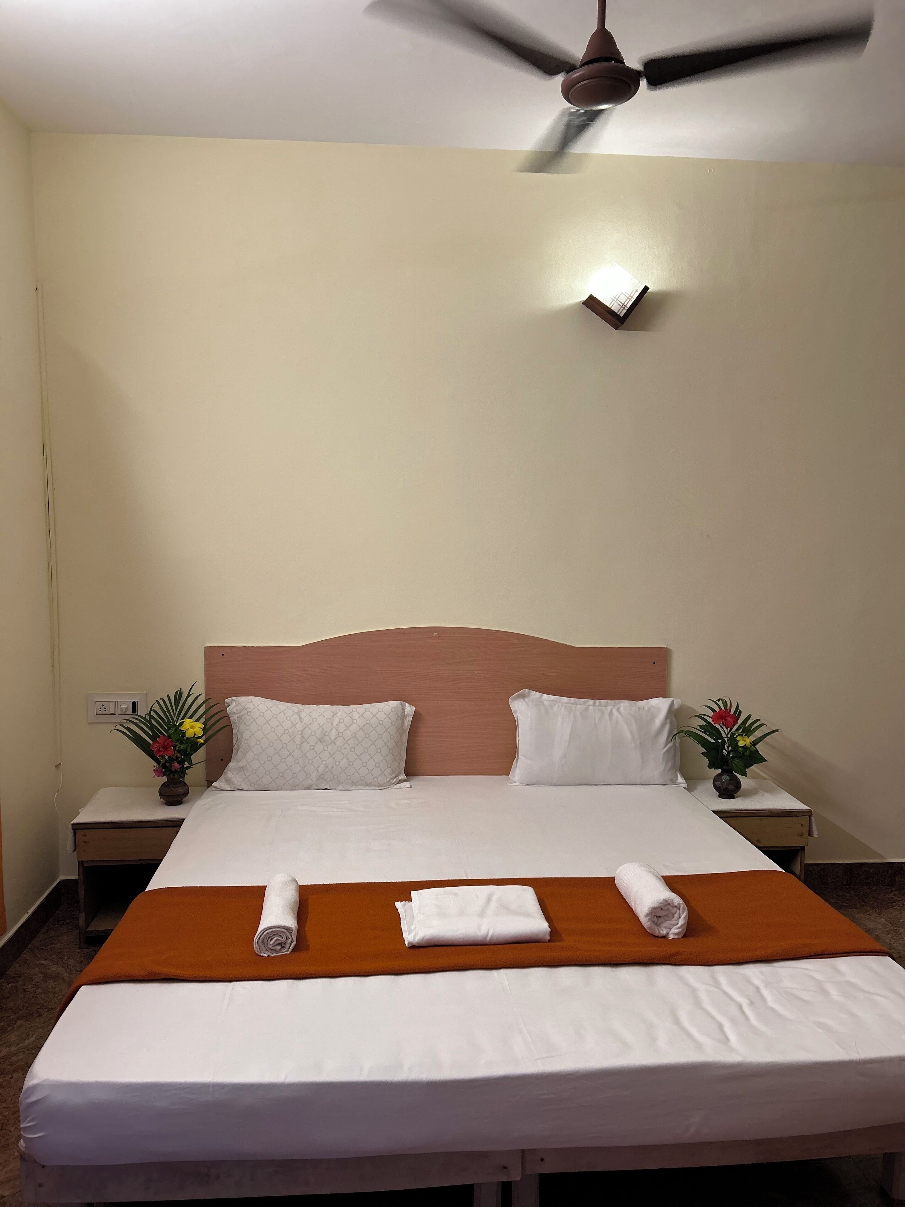 Una cama bien hecha con sábanas blancas, dos almohadas, una manta marrón y dos toallas enrolladas. Hay dos mesitas de noche con jarrones de flores, un ventilador de techo y un enchufe de pared.