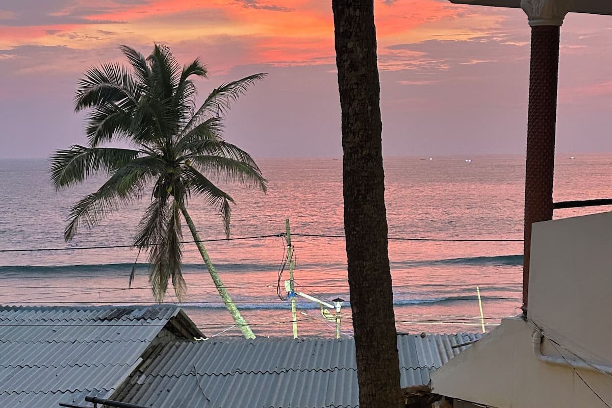 Blick auf die Küste bei Sonnenuntergang, mit Palmen und einem Gebäude mit Wellblechdächern.
