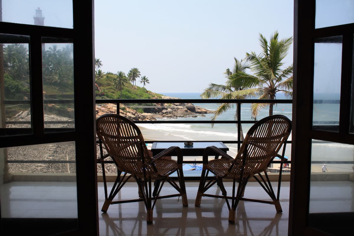 Ein Balkon mit zwei Korbstühlen und einem kleinen Tisch, der Blick auf einen Strand mit Palmen und einer felsigen Küste freigibt.