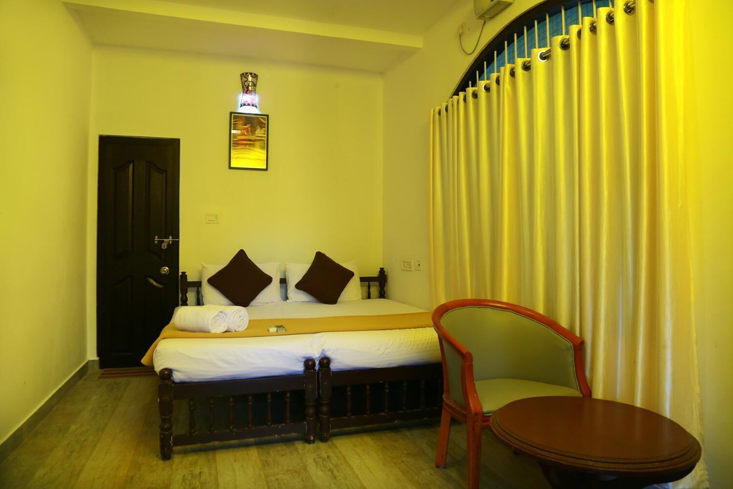 A hotel room with a bed, a chair, a small table, a door, and a wall-mounted light fixture.