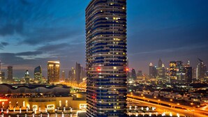 A high-rise building with a distinctive circular top, surrounded by a cityscape with illuminated buildings and a busy highway. - DAMAC Maison Mall Street (Dubai)