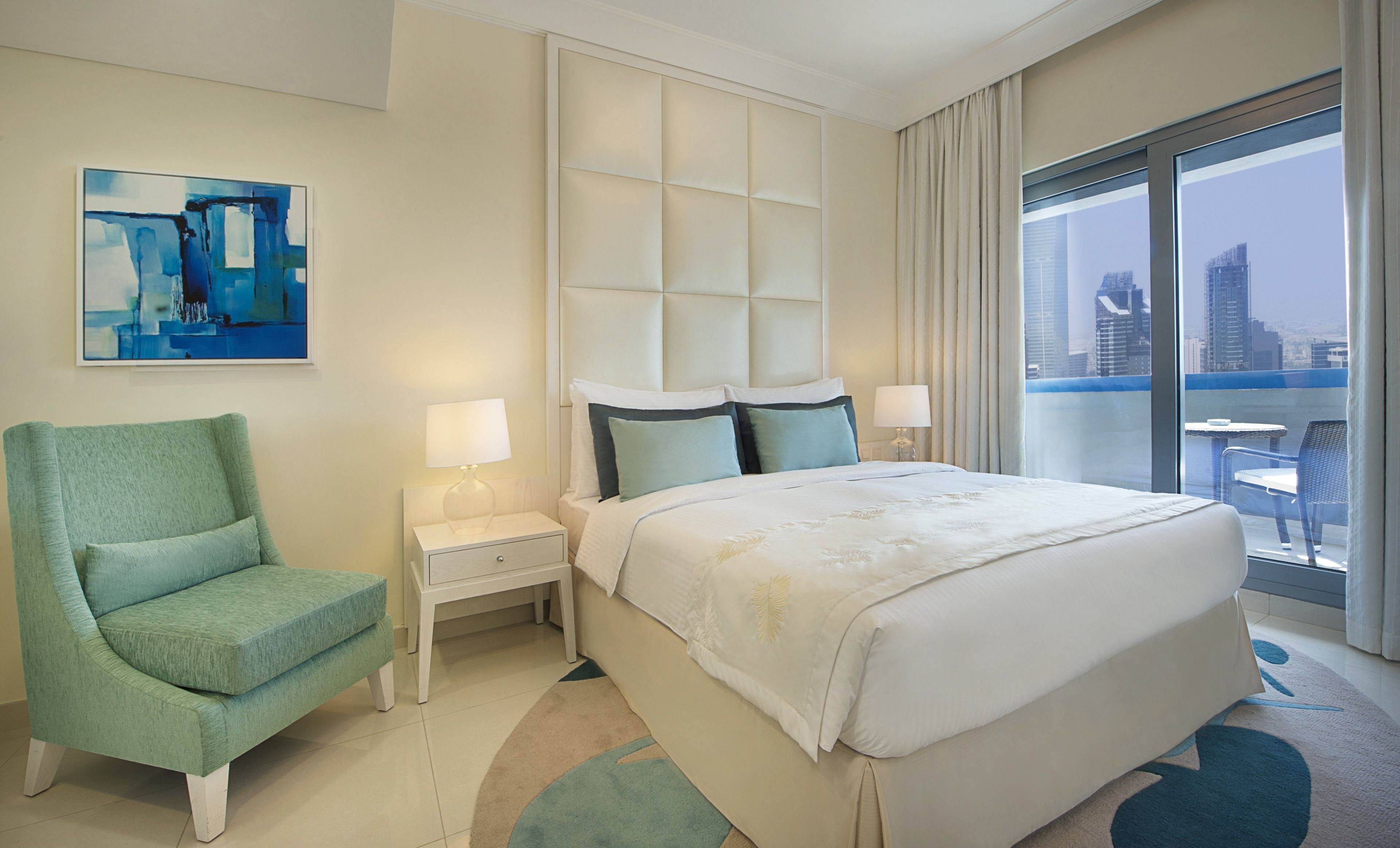 Habitación de hotel con una cama grande, una mesita de noche, una silla, un cuadro y un balcón con vistas a la ciudad.