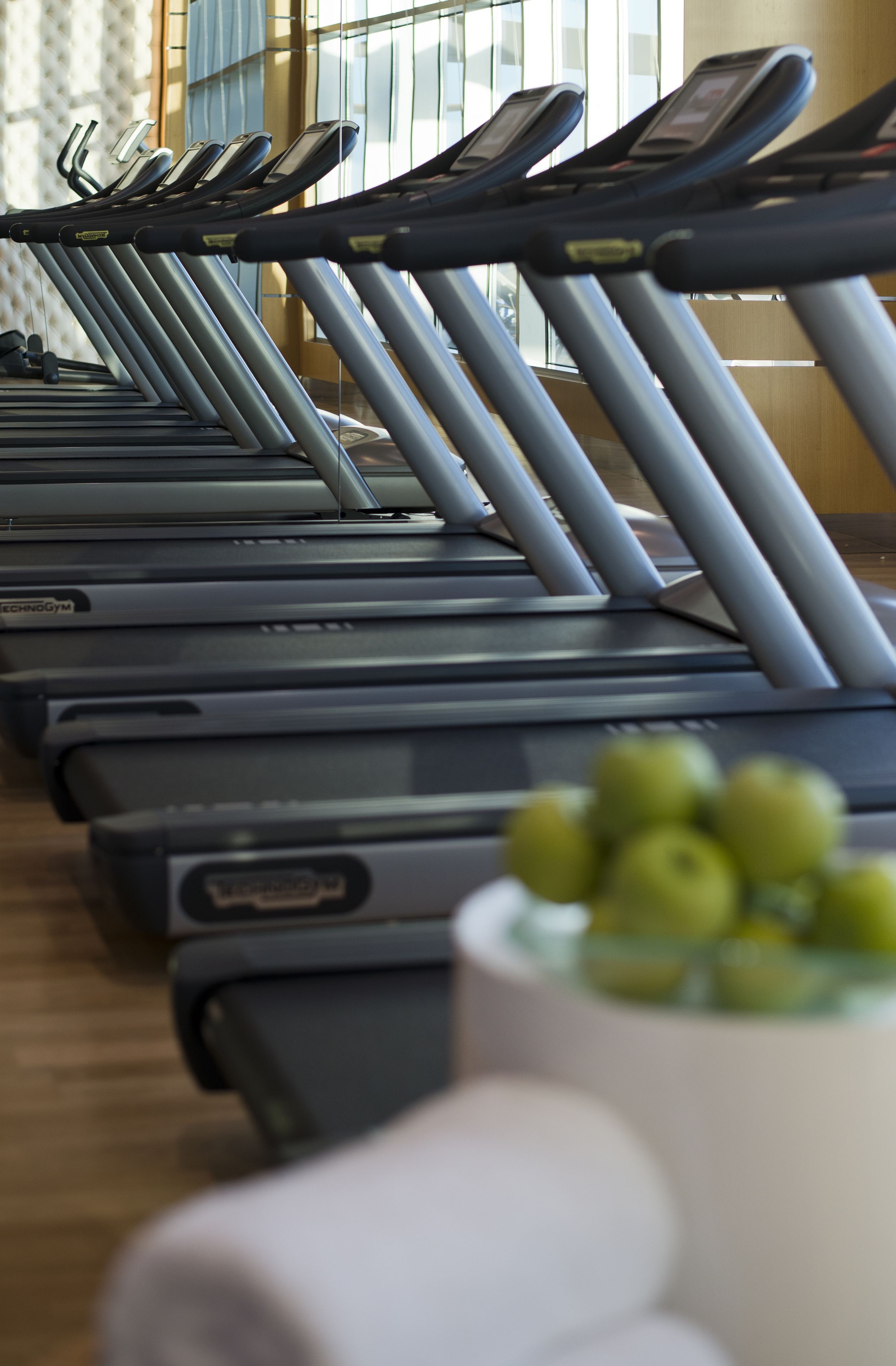A row of treadmills in a gym with a wooden floor and a window in the background.