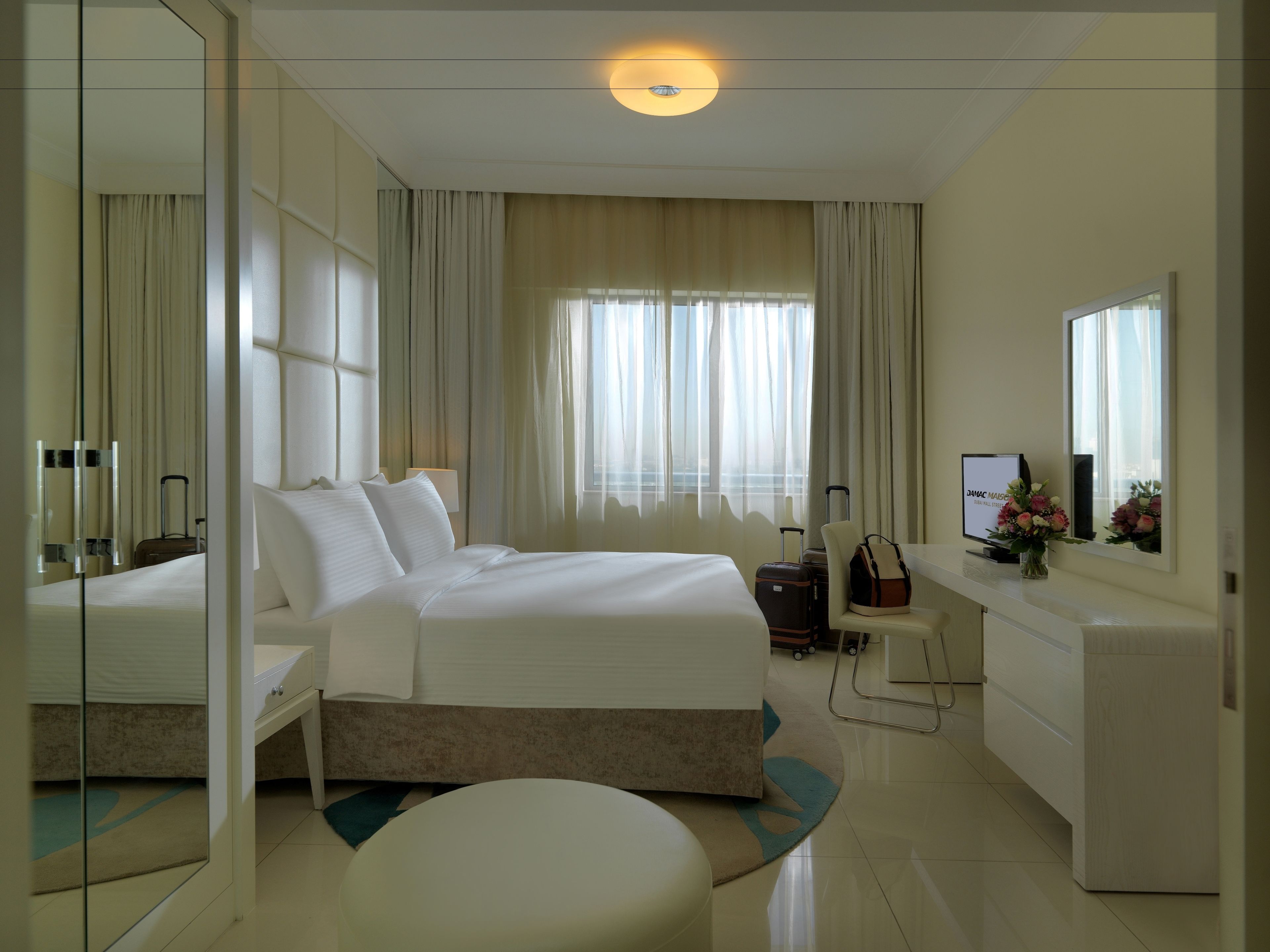 Una habitación de hotel con una cama grande, un escritorio con una computadora, una silla, una mesita con un jarrón de flores y una maleta.