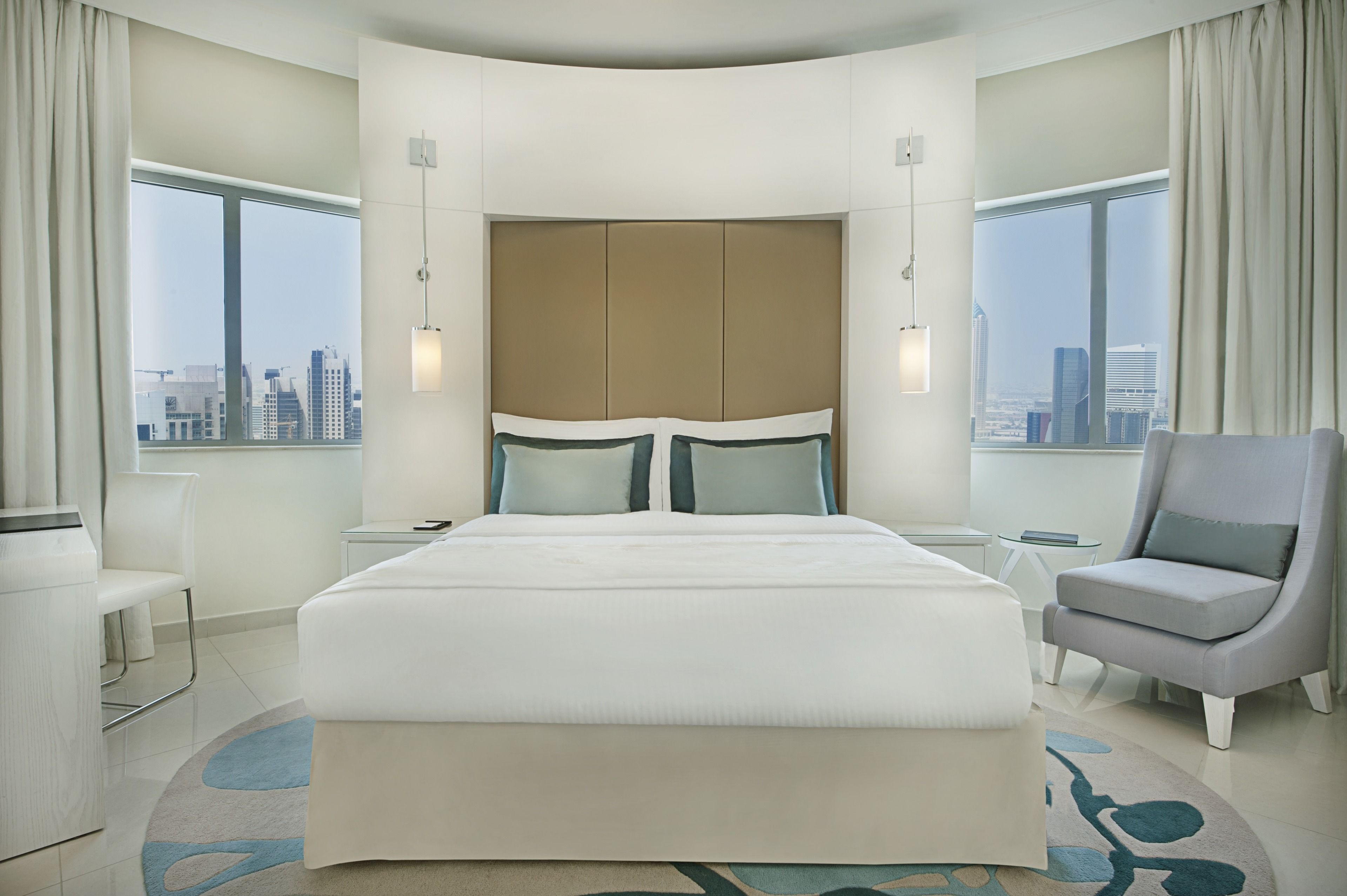 Ein modernes Hotelzimmer mit einem großen Bett, einer Sitzecke mit einem Sessel und Blick auf die Stadt durch große Fenster.