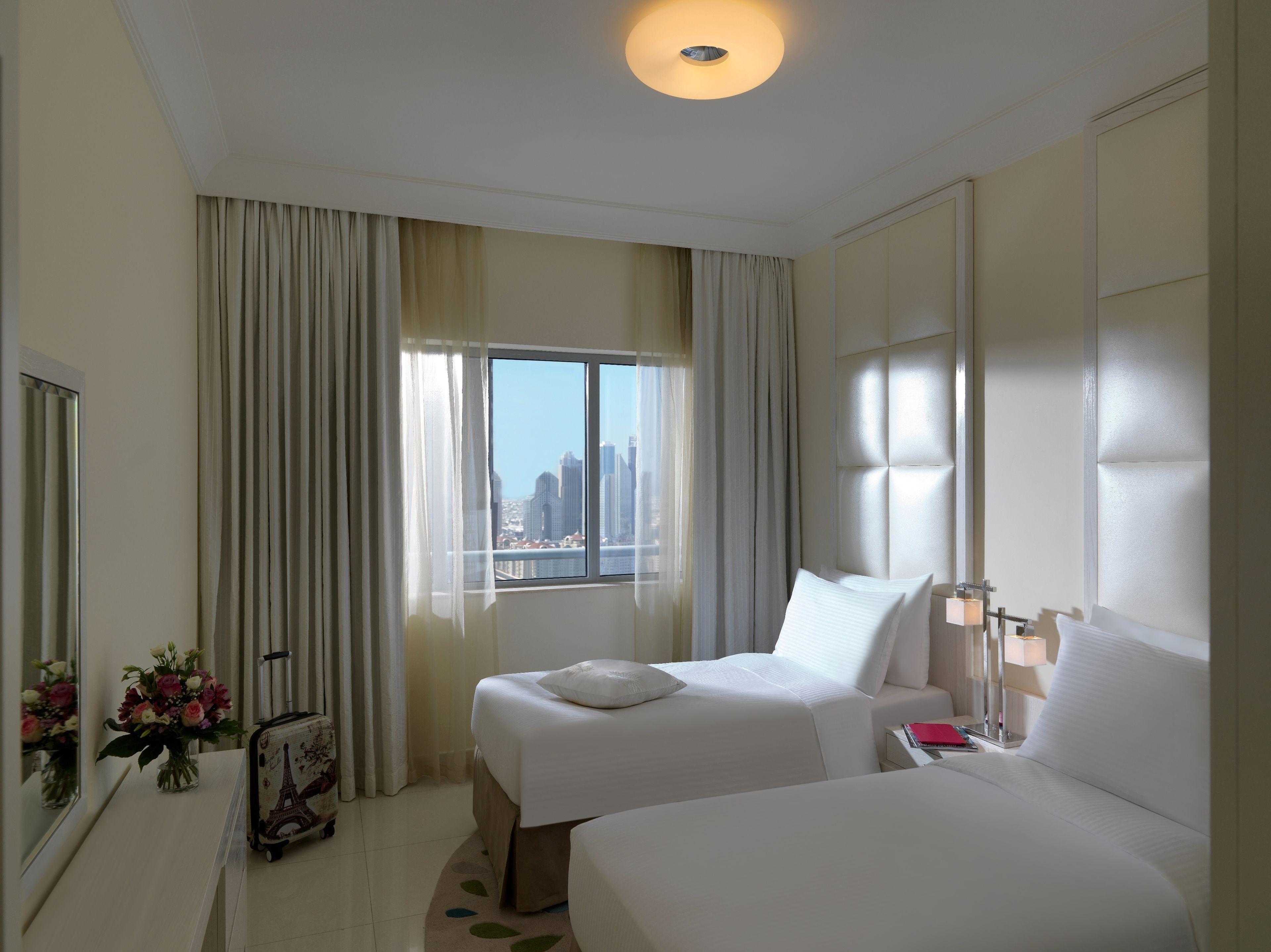 A hotel room with two beds, a window with a city view, a mirror, and a vase of flowers.