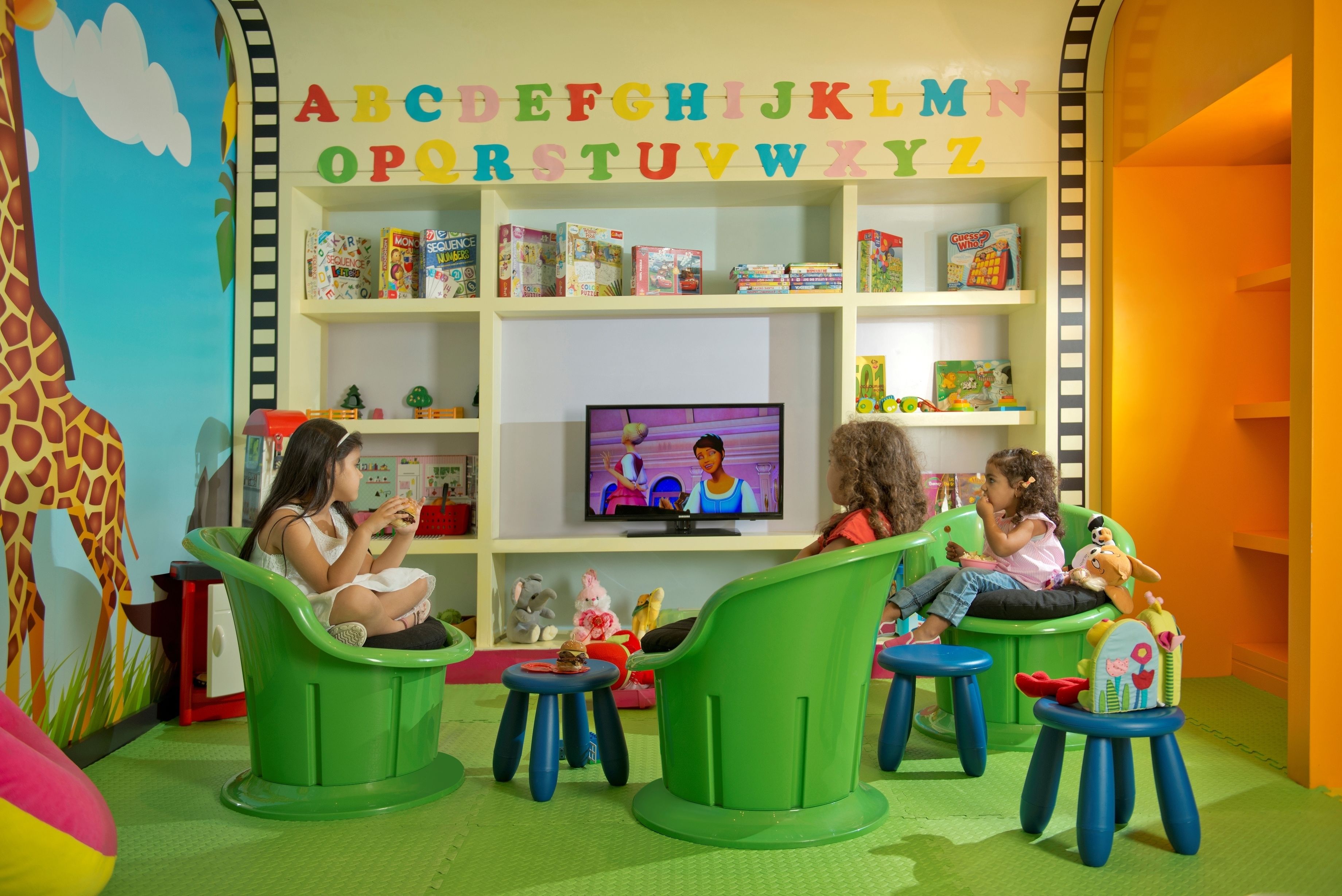 A children's play area with green chairs, a television, and alphabet-themed shelves.