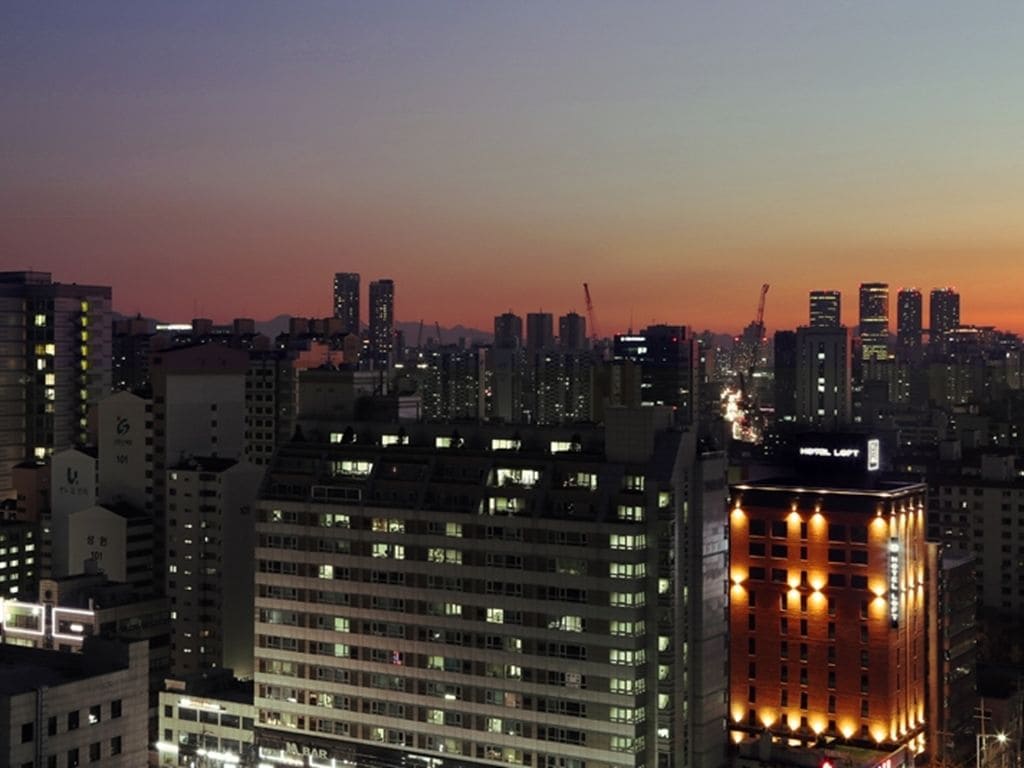 A cityscape at dusk with illuminated buildings, including a hotel with a distinctive facade and a bar.