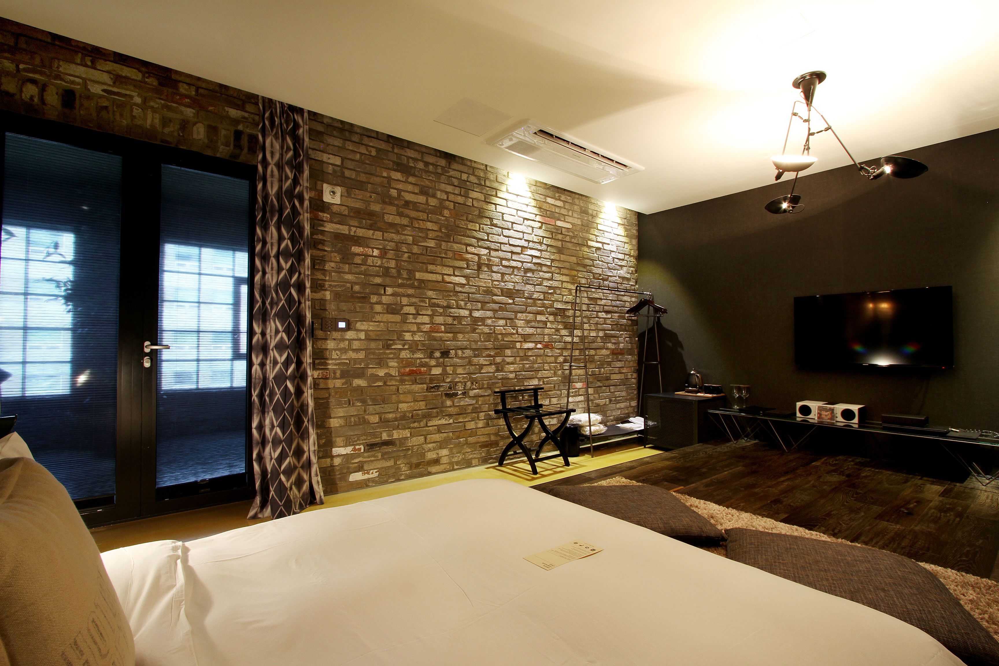 A hotel room with a brick wall, a bed with white linens, a television, and a desk with speakers.
