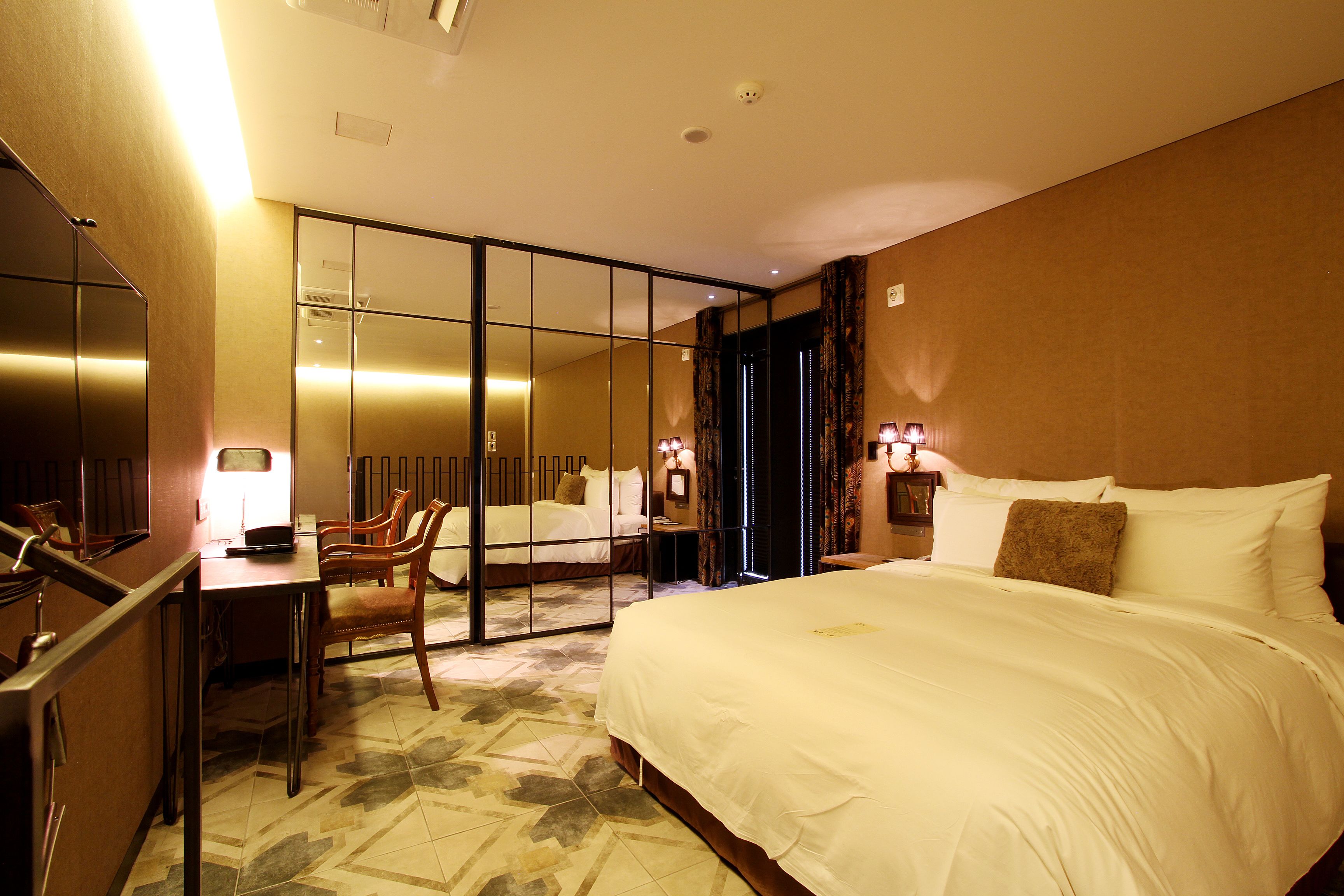 A hotel room with a large bed, a desk, and a chair. There is a mirror on the wall and a window with curtains.