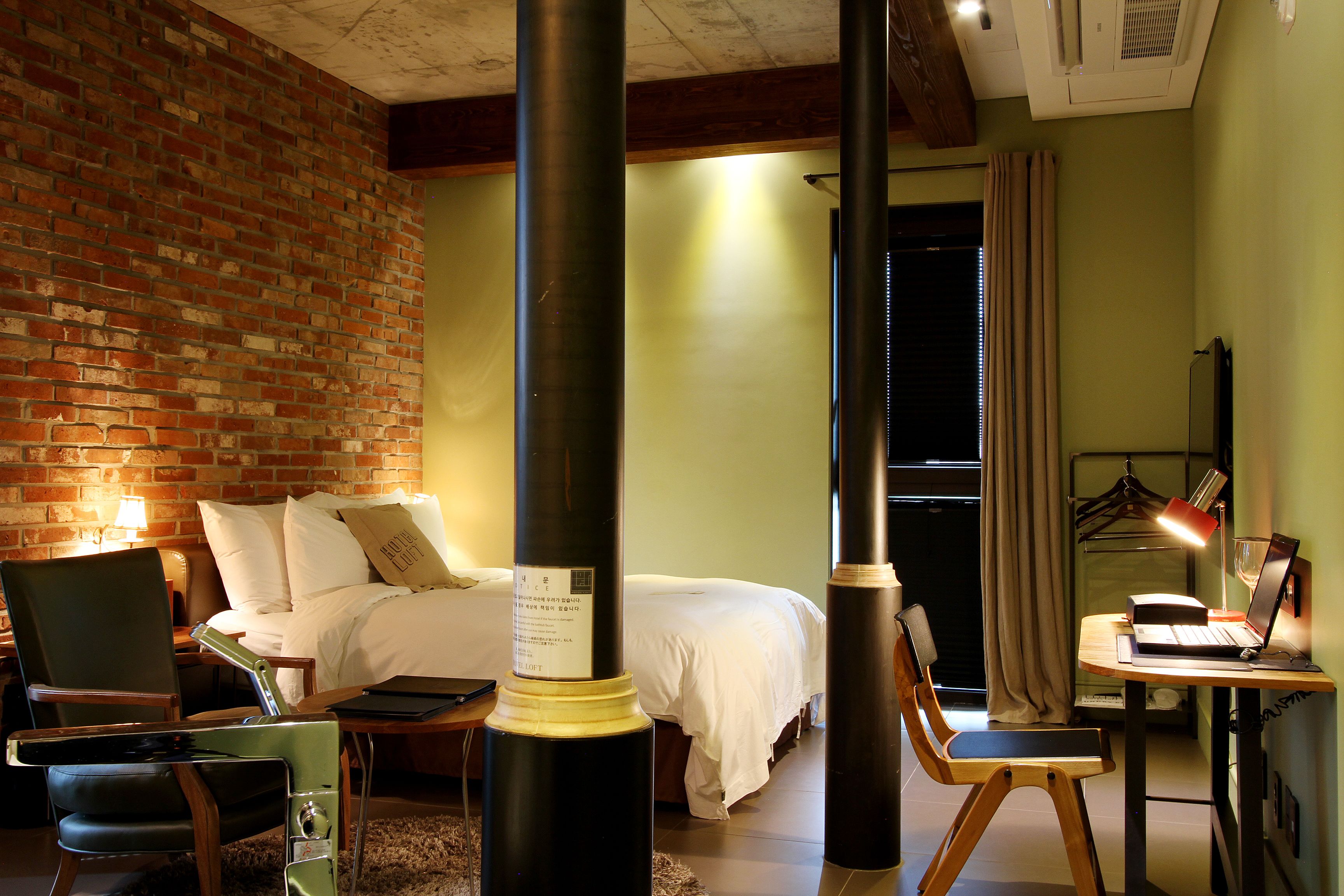 A hotel room with a brick wall, a bed with white bedding, a desk with a laptop, and a chair.