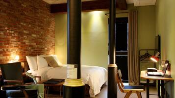 A hotel room with a brick wall, a bed with white bedding, a desk with a laptop, and a chair.