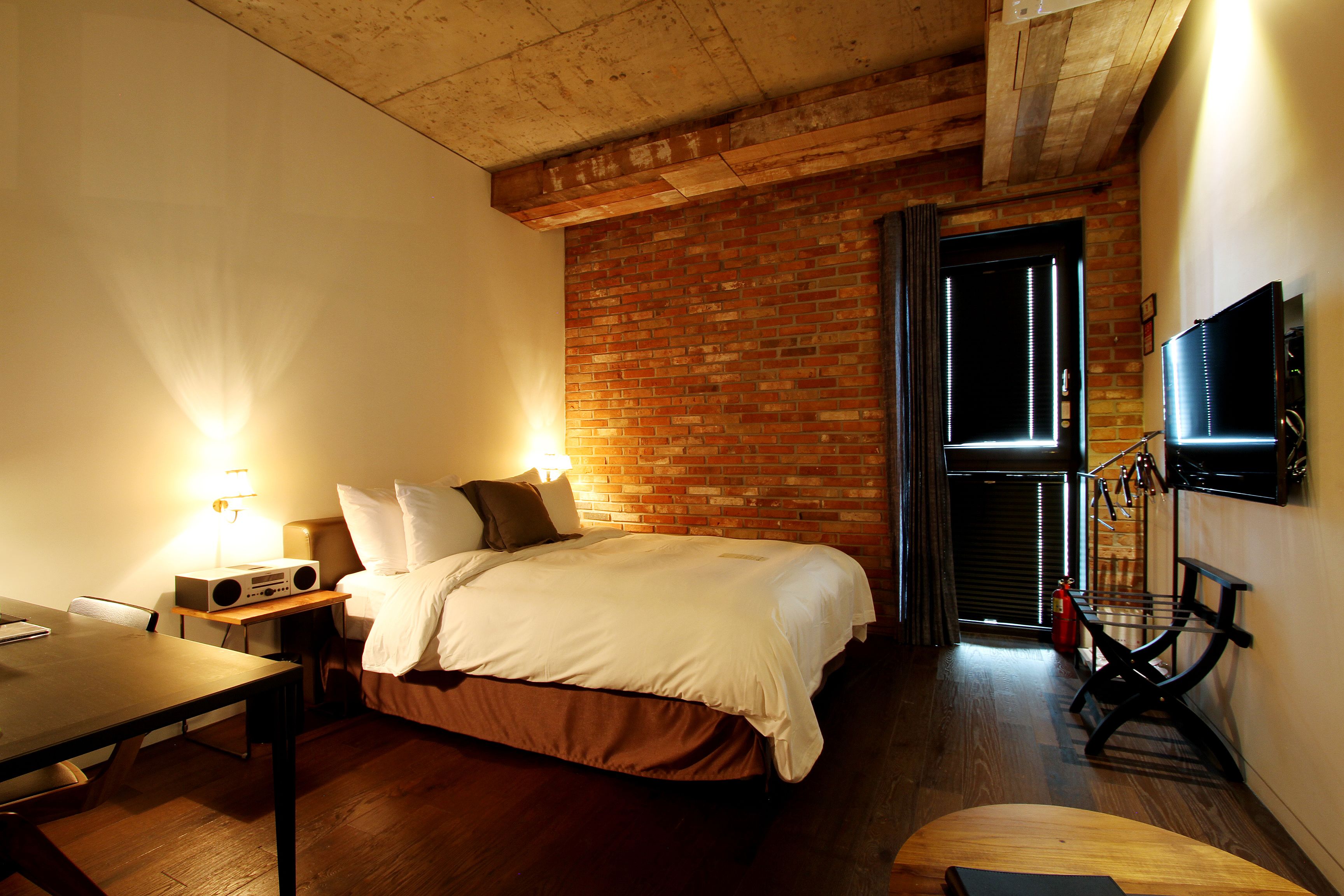 A hotel room with a brick wall, a bed with white bedding, a desk with a lamp, and a television mounted on the wall.