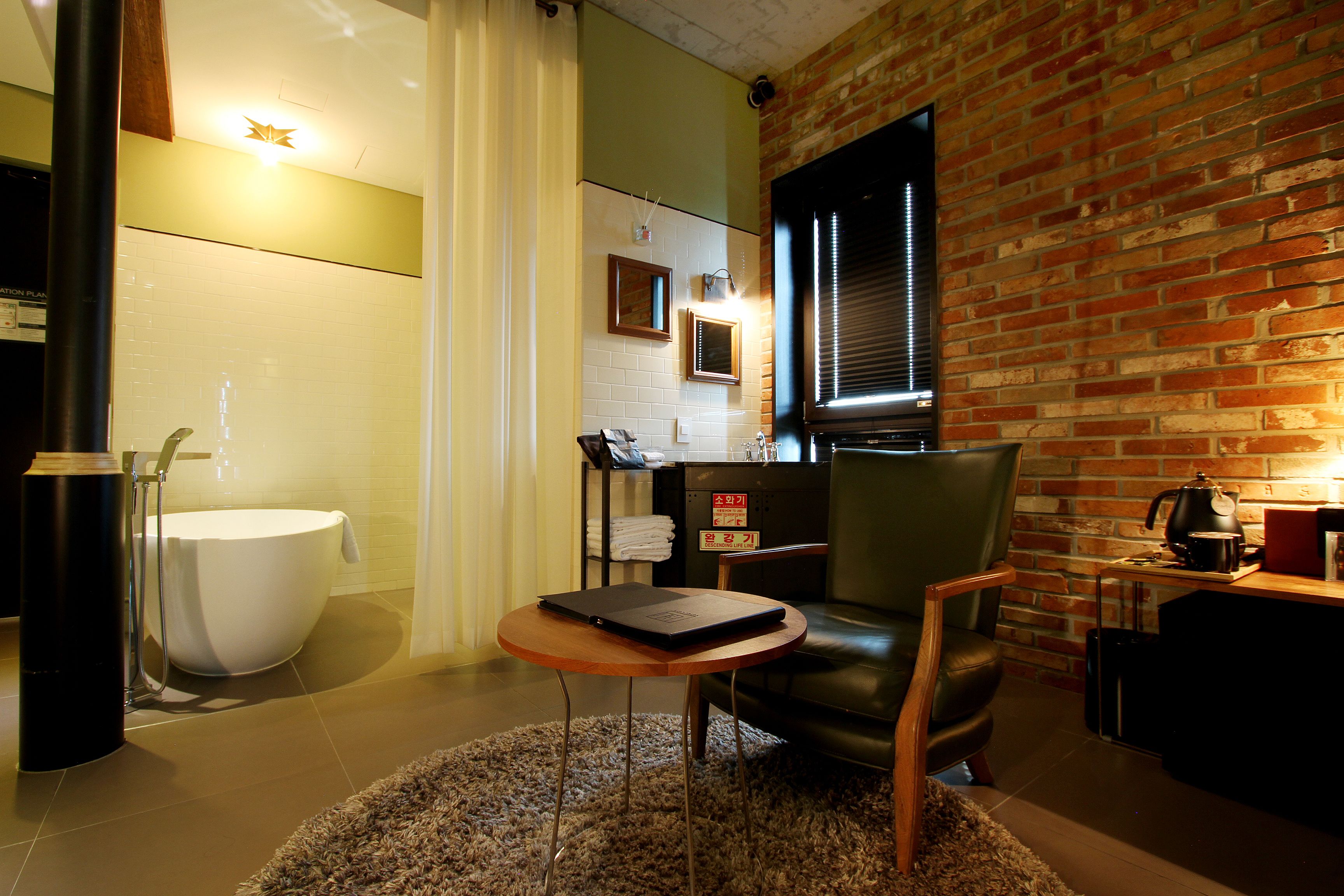 A modern bathroom with a bathtub, a sink, and a shower. There is a chair and a table with a laptop. The room has a brick wall and a window with blinds.