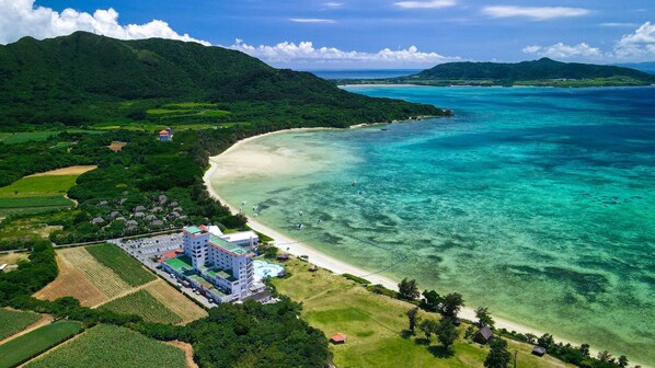 Vista aérea de una zona costera con un gran complejo de edificios cerca de una playa, rodeada de exuberante vegetación y aguas azul cristalino.