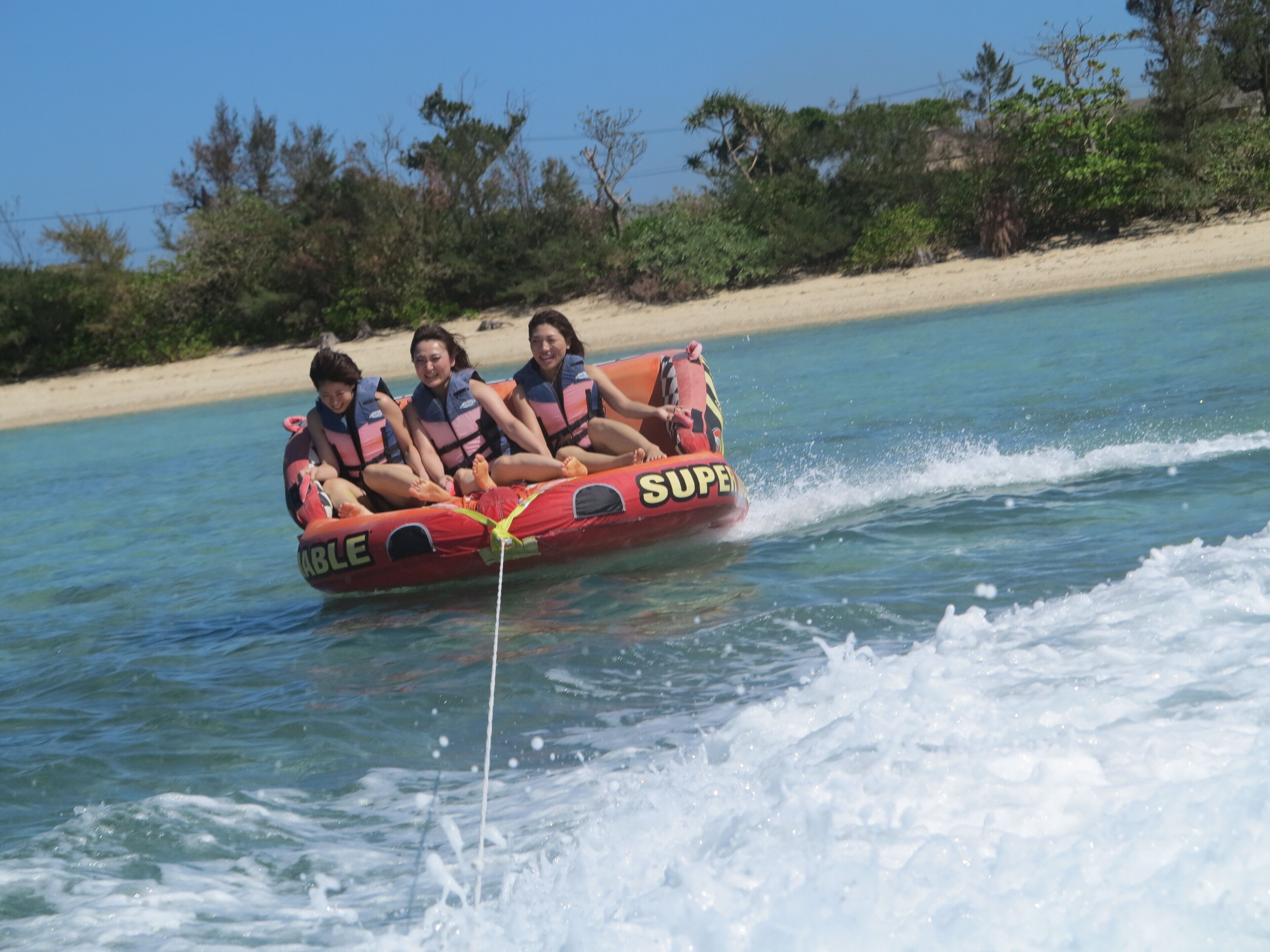 A watercraft with three people wearing life jackets, moving on a body of water with a sandy beach and greenery in the background.