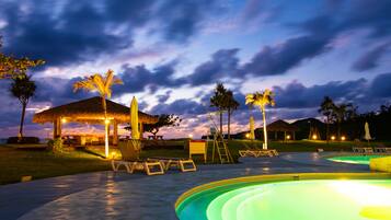 A pool area with lounge chairs, a thatched-roof structure, and palm trees.