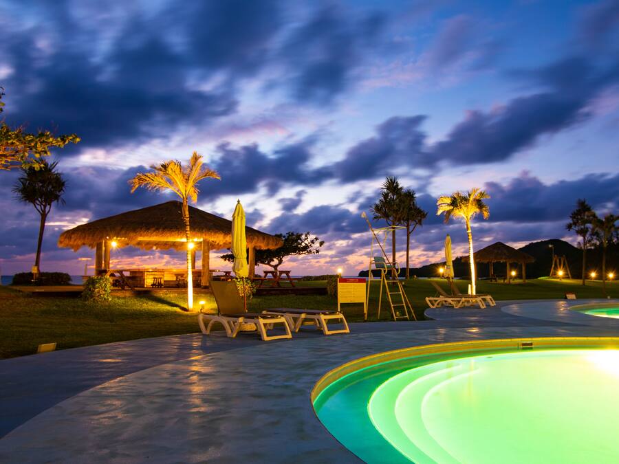 A pool area with lounge chairs, a thatched-roof structure, and palm trees.