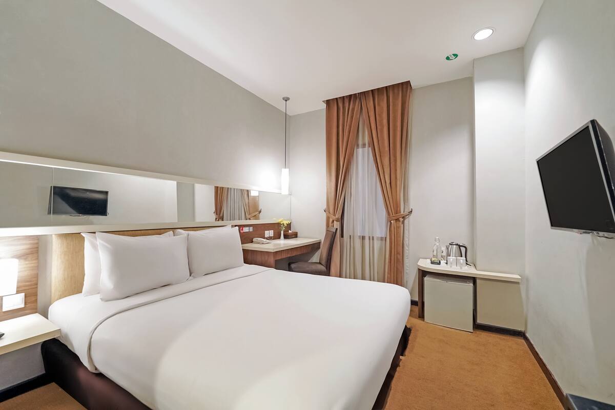 Ein Hotelzimmer mit einem großen Bett, einem Schreibtisch mit Stuhl, einem kleinen Tisch mit Lampe, einem an der Wand befestigten Fernseher und einem Fenster mit Vorhängen.