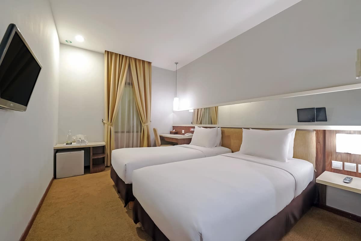 Ein Hotelzimmer mit zwei Betten, einem Schreibtisch, einer kleinen Kühlschrank, einem Fernseher und einem Nachttisch mit Lampe.
