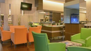 Un hall d’hôtel moderne avec un plancher en bois, des sièges verts et orange, un comptoir de réception et un téléviseur fixé au mur.