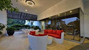 A modern outdoor lounge area with a white wicker sofa set, a glass wall, and a ceiling light fixture. - Amaroossa Royal Bogor (Bogor)