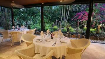 An outdoor dining area with wicker chairs and round tables, surrounded by lush greenery and tropical plants.