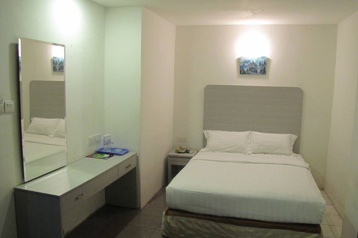 A hotel room with a bed, a bedside table, a desk, a mirror, and a small painting on the wall.