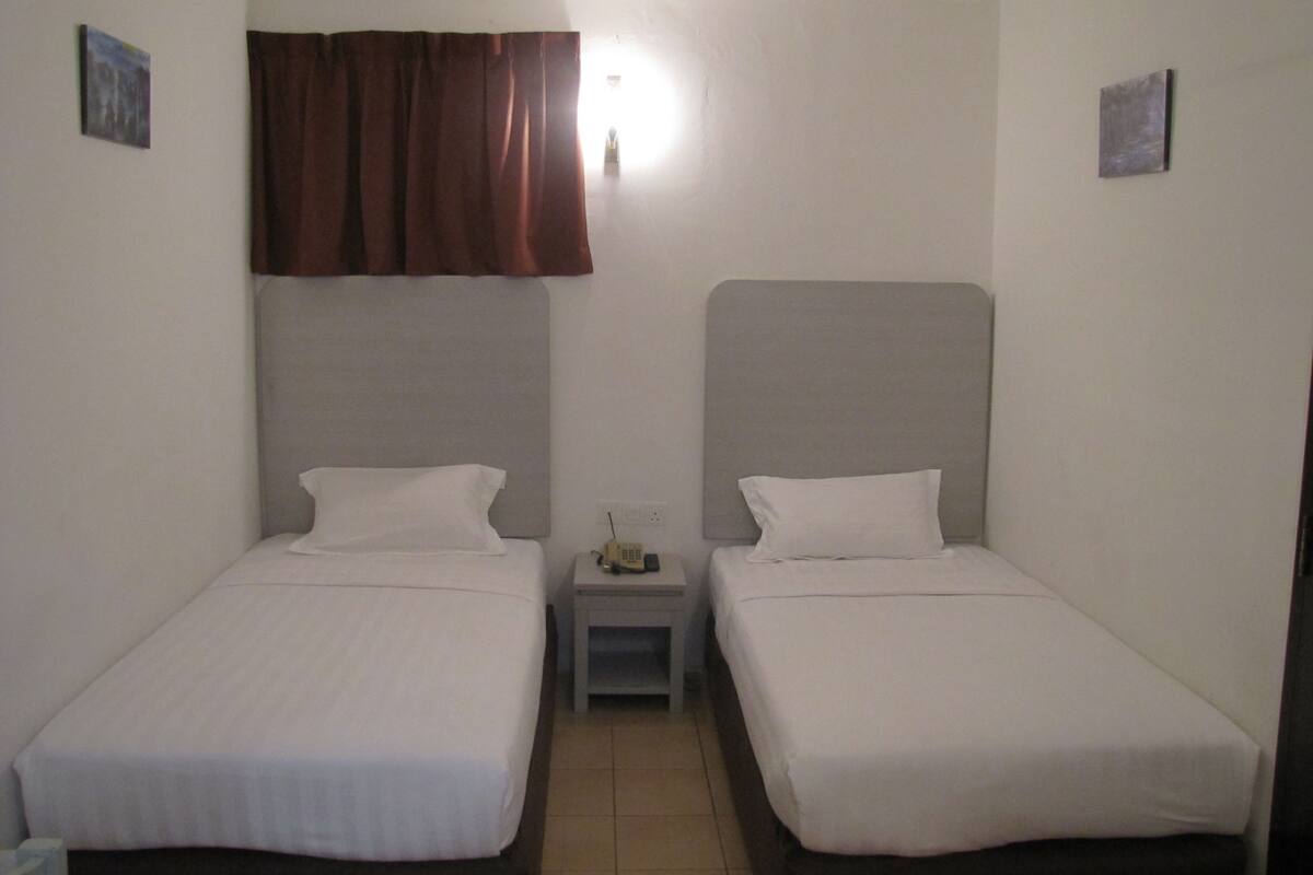 A small, clean room with two single beds, a nightstand with a lamp, and a window with a curtain.