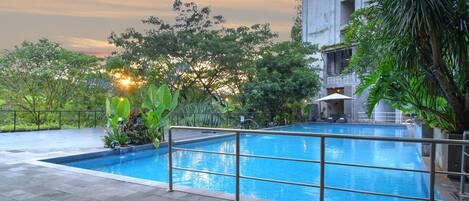 An outdoor swimming pool with a metal railing, surrounded by greenery and a building in the background.