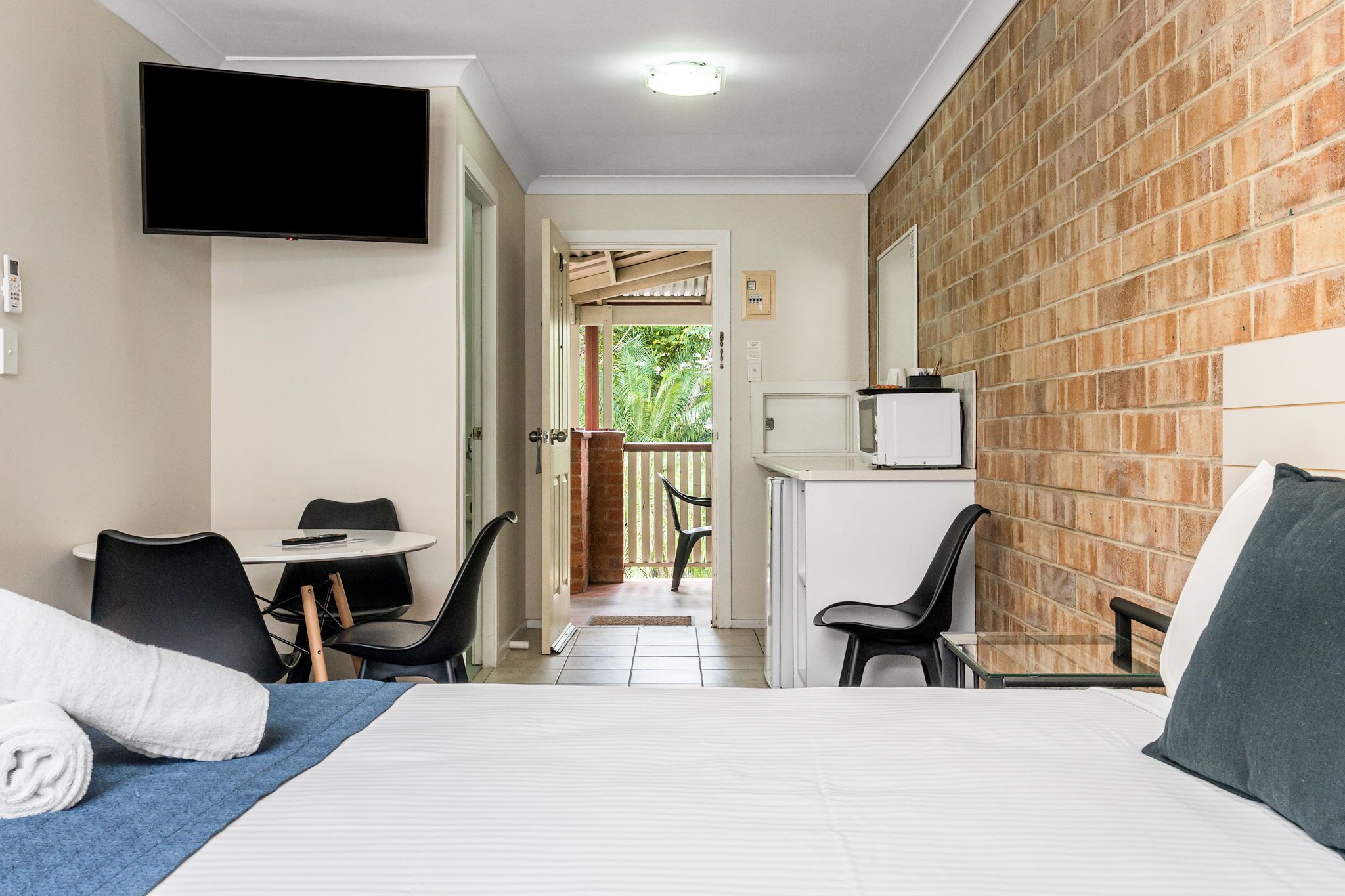 Habitación de hotel con una cama, televisor montado en la pared, un pequeño escritorio con una silla y un balcón con vistas a zonas verdes.