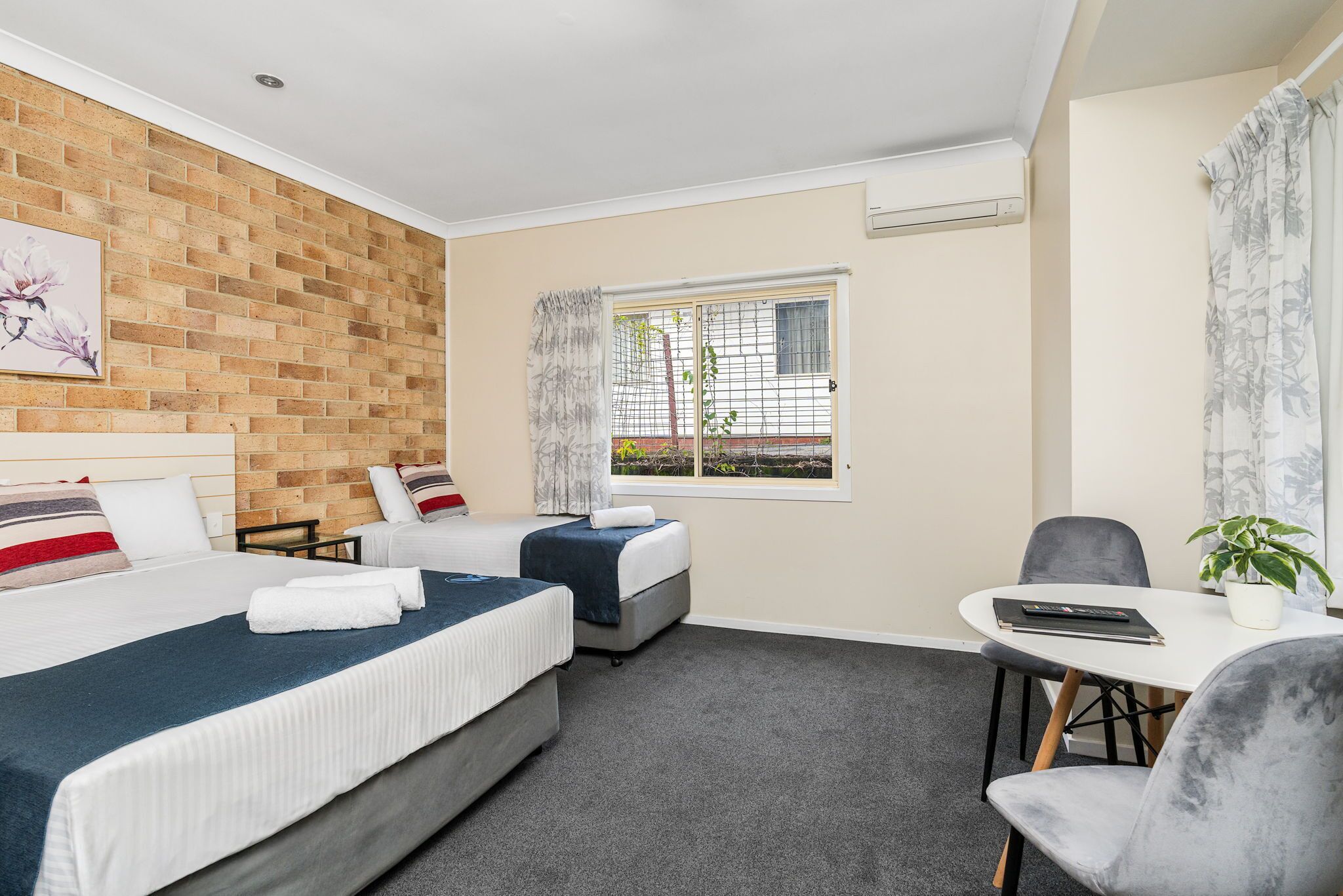 A hotel room with two beds, a small table with a chair, a potted plant, and a window with curtains.